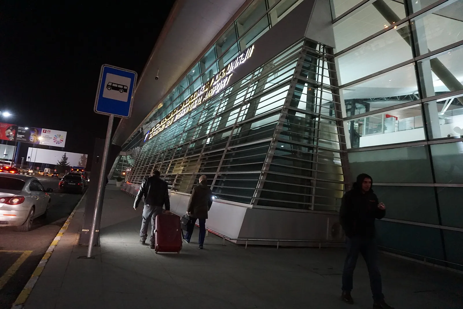 Voyageurs avec une valise devant la façade vitrée du terminal de l'aéroport international de Tbilissi, de nuit