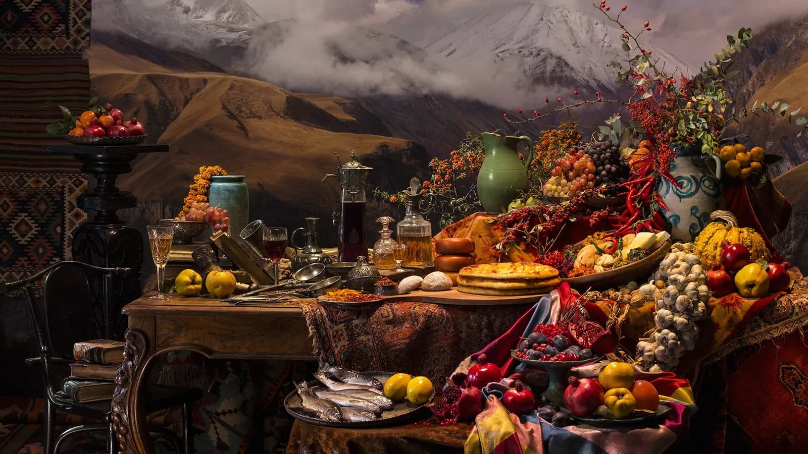 Nature morte d'une table de fête géorgienne : pains, fromages, grenades, coings, ail, piments et carafes de vin devant un décor de montagnes enneigées