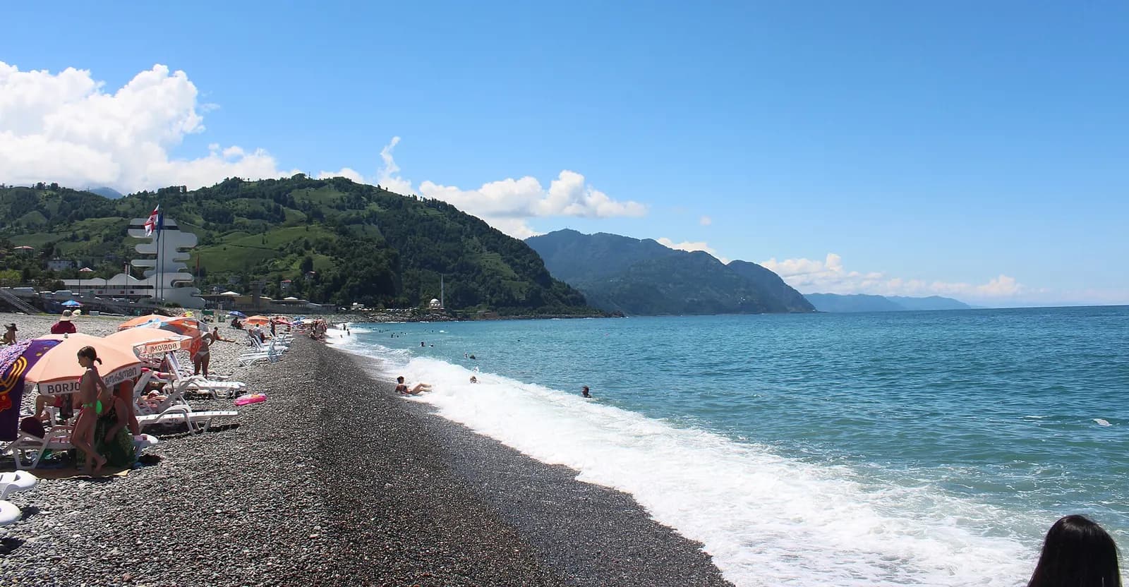 Plage de galets de Sarpi, avec parasols, vagues et collines vertes