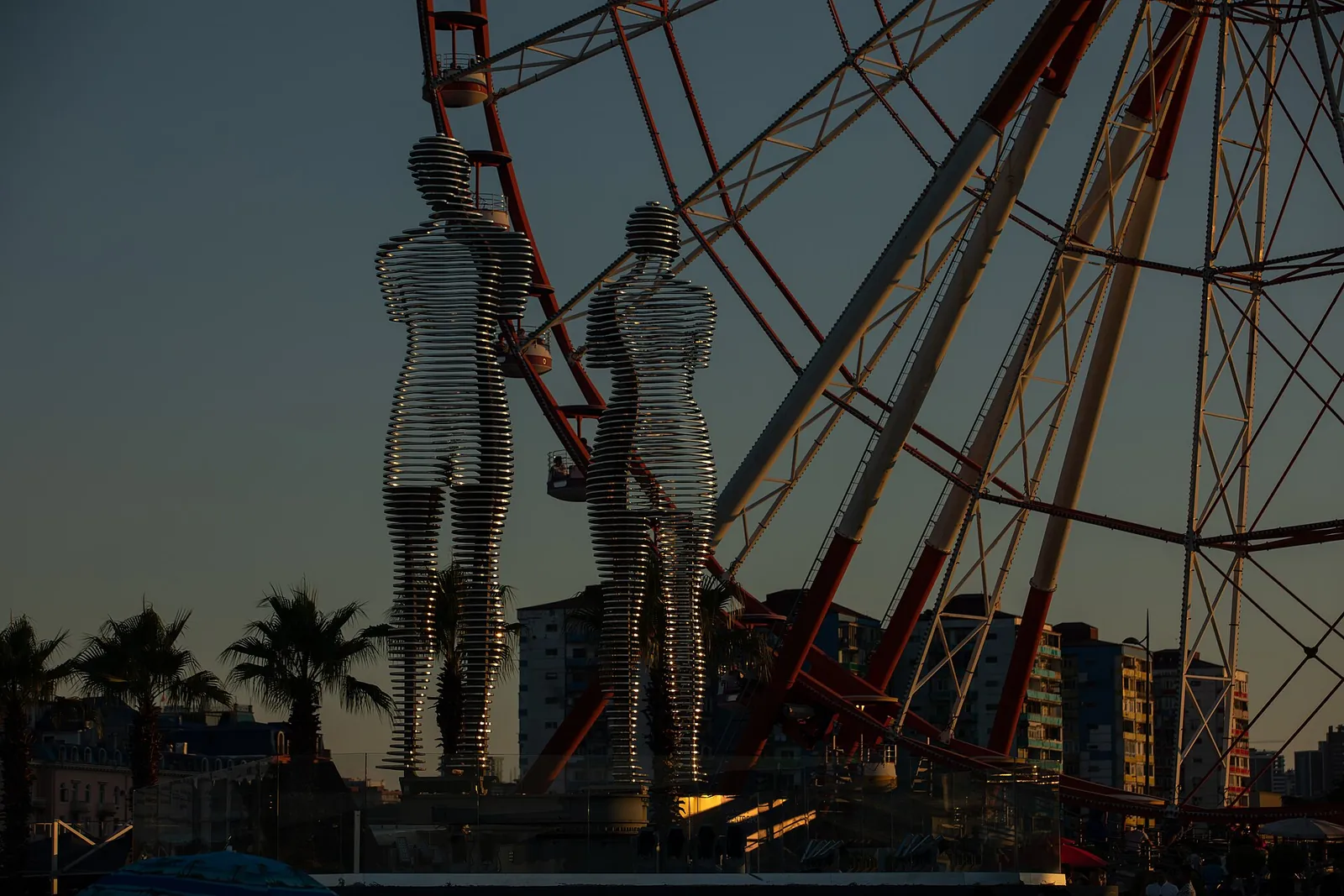 Statue mobile Ali et Nino, deux silhouettes d'anneaux métalliques devant la grande roue du boulevard de Batoumi au crépuscule