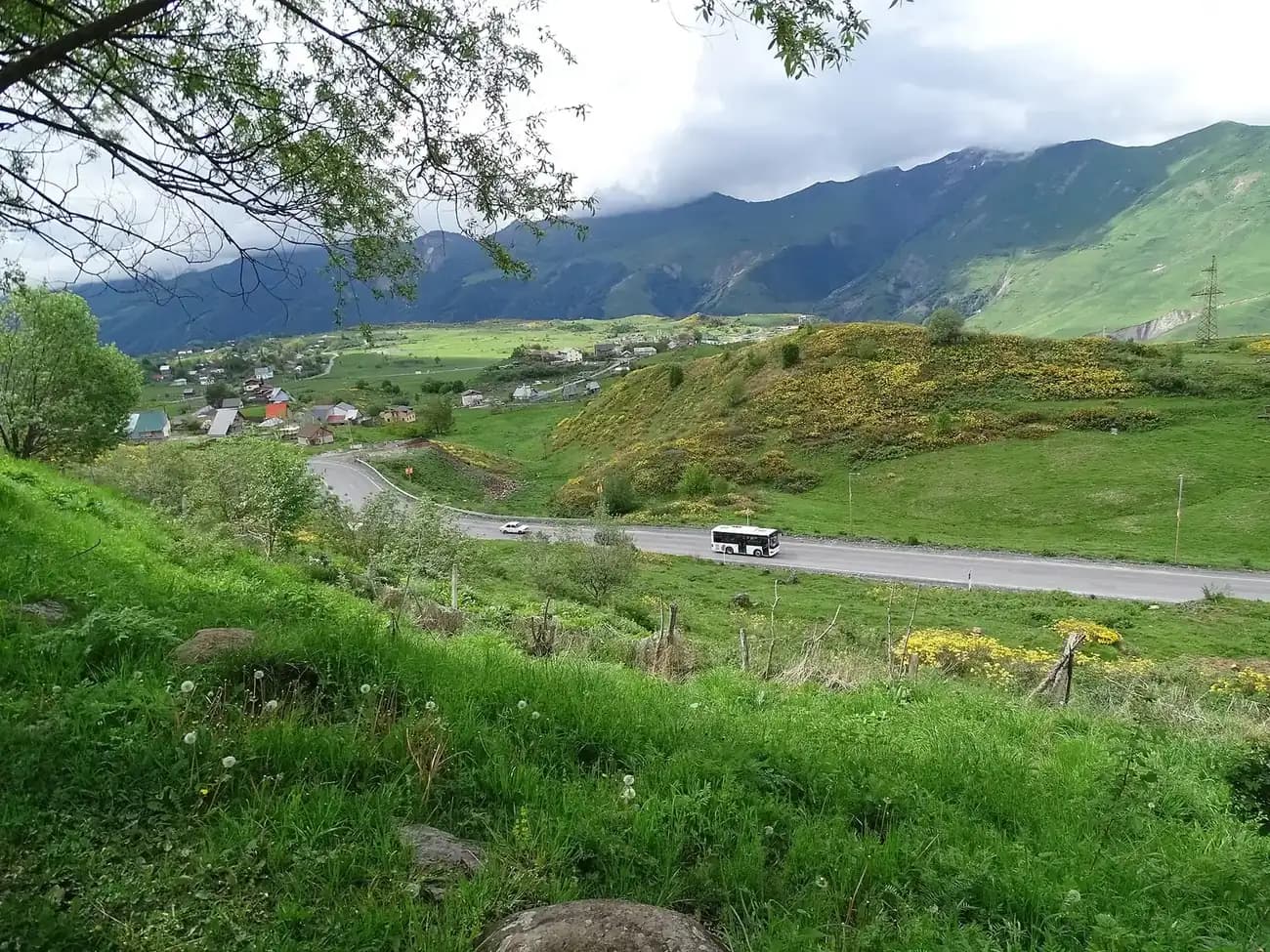 Route militaire géorgienne en lacets près de Gudauri au printemps, avec un bus, des genêts en fleurs et le village en contrebas