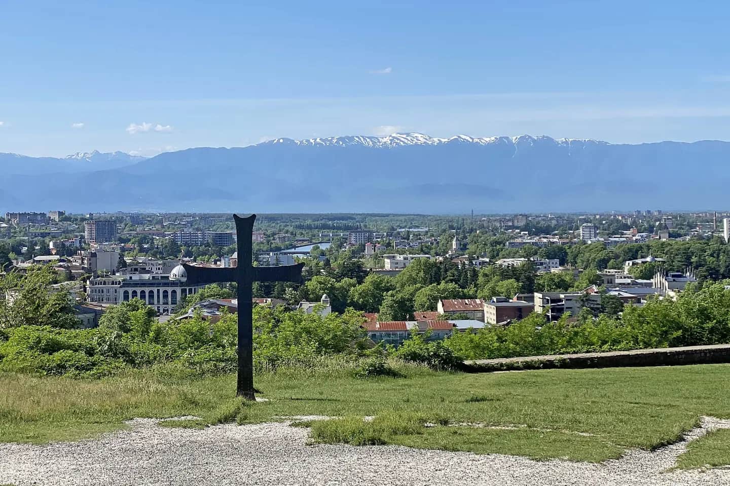Vue sur Koutaïssi depuis la colline de Bagrati, avec une grande croix en métal au premier plan et la chaîne de montagnes enneigée à l’horizon