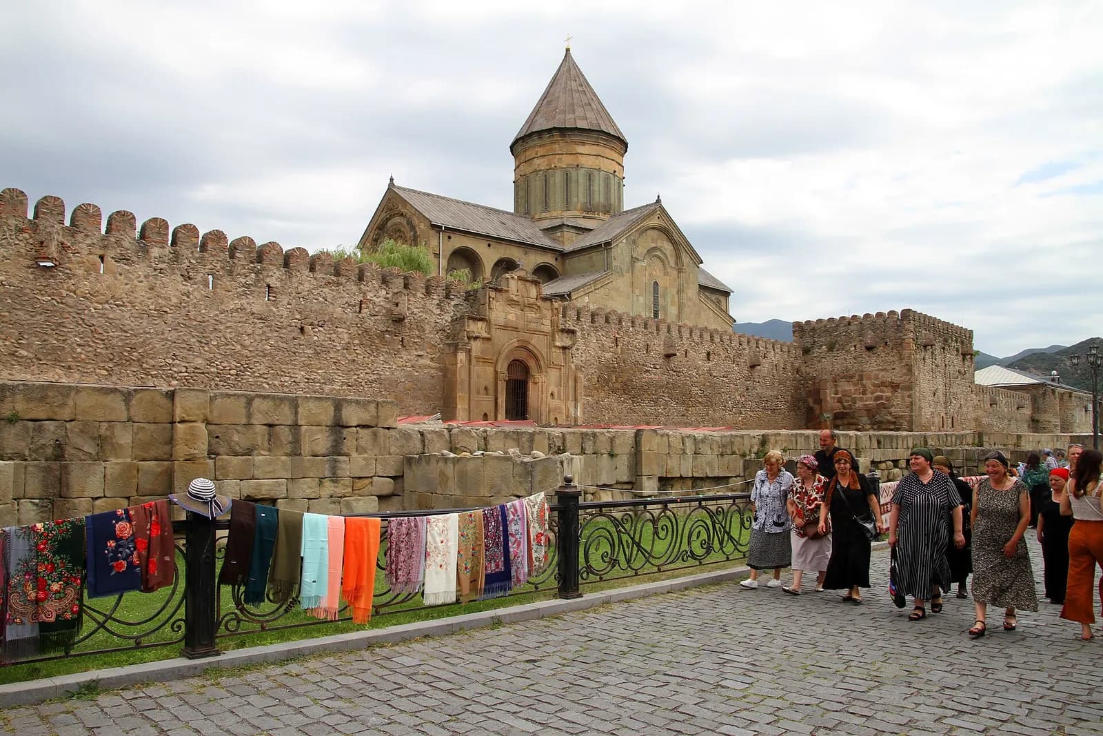 Remparts crénelés qui entourent la cathédrale de Svetitskhoveli à Mtskheta, avec des écharpes en vente le long de la grille et des visiteurs sur les pavés