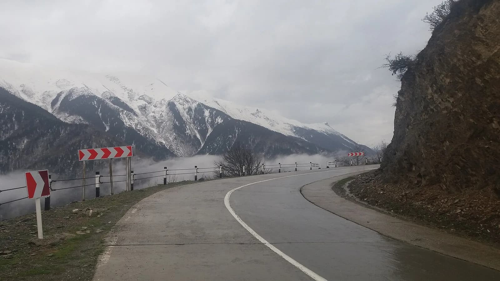 Virage de la route de Mestia en Svanétie, bordée de balises rouges et blanches, face aux montagnes enneigées du Caucase dans la brume