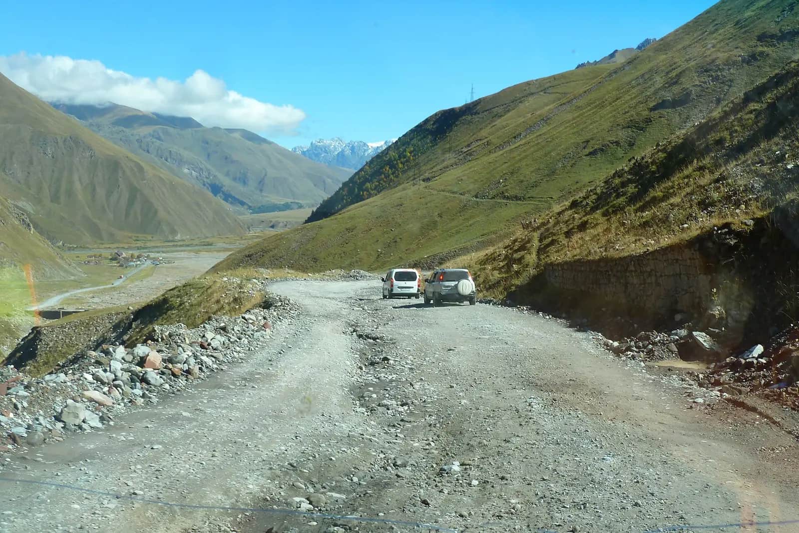 Deux voitures sur une piste de gravier en lacet au nord du col de Jvari, sur la route militaire géorgienne, avec la vallée vers Stepantsminda et les sommets du Grand Caucase au fond