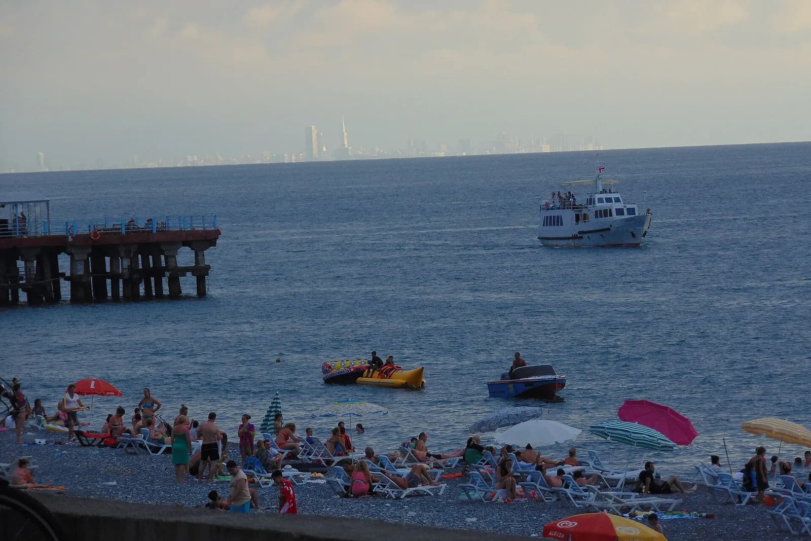 Plage de galets de Kobuleti en soirée d'été avec baigneurs, ponton et bateau, la skyline de Batoumi au loin