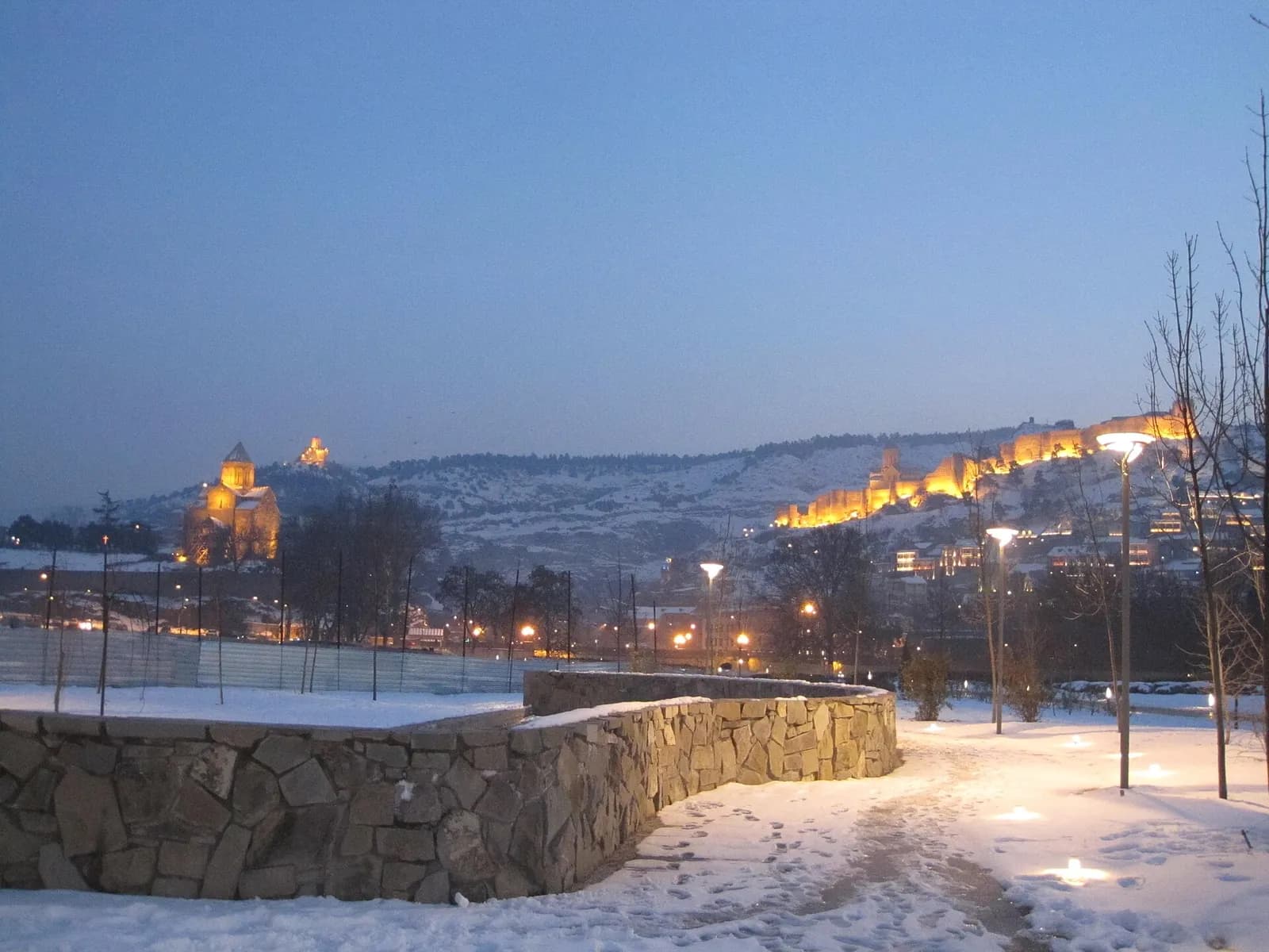 Parc Rike enneigé à Tbilissi au crépuscule, avec l'église de Metekhi et la forteresse de Narikala illuminées sur la colline